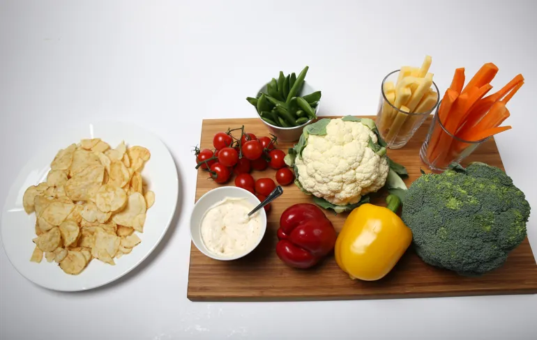 Et brett med diverse grønnsaker; paprika brokkoli, gulrøtter, næpe, sukkererter og tomat. En skål med dip og en skål siden av med potetgull.