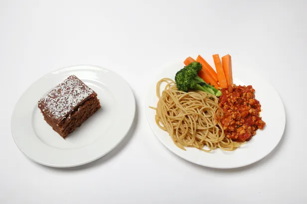 Et fat med et sjokoladekakestykke og et fat med spagetti, kjøttsaus, gulrøtter i staver og en brokkolibusk.