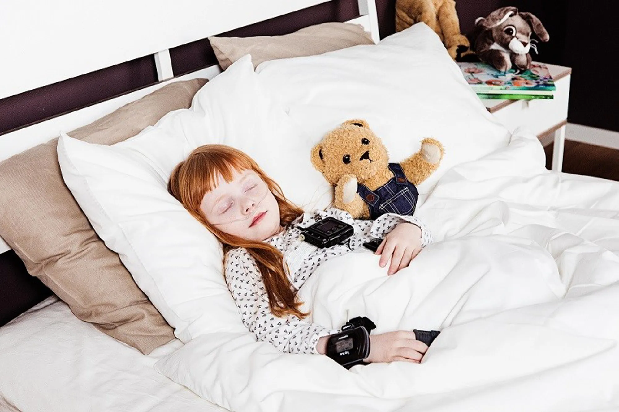 Barn ligger i seng og sover, med bamse ved siden av seg. Måleapparater på seg. Foto.