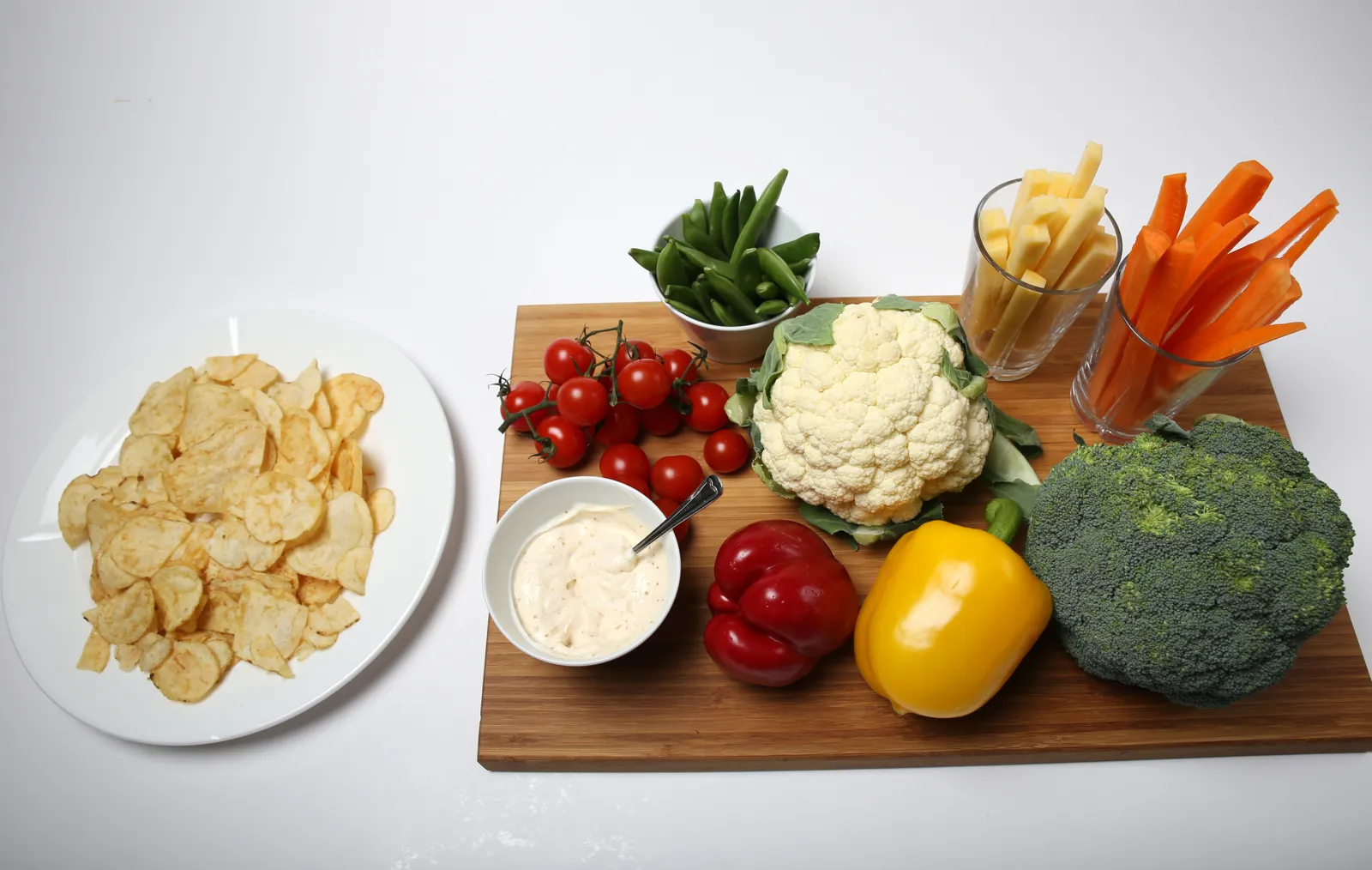 Et brett med diverse grønnsaker; paprika brokkoli, gulrøtter, nepe, sukkererter og tomat. En skål med dip og en skål siden av med potetgull.