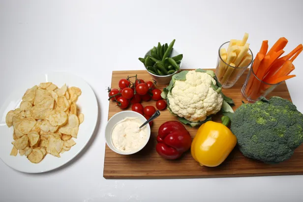 Et brett med diverse grønnsaker; paprika brokkoli, gulrøtter, nepe, sukkererter og tomat. En skål med dip og en skål siden av med potetgull.
