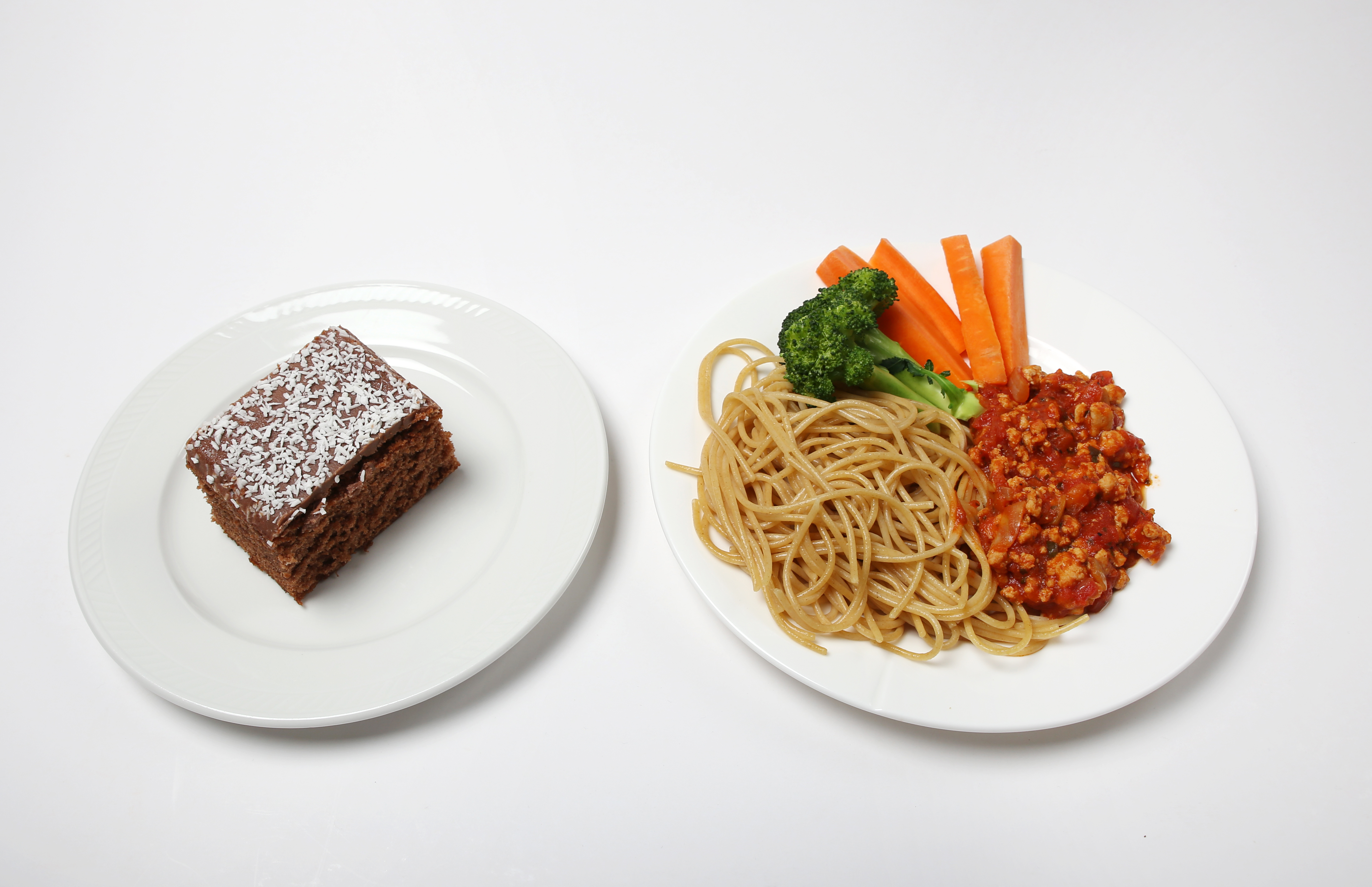 Et fat med et sjokoladekakestykke og et fat med spagetti, kjøttsaus, gulrøtter i staver og en brokkolibusk.