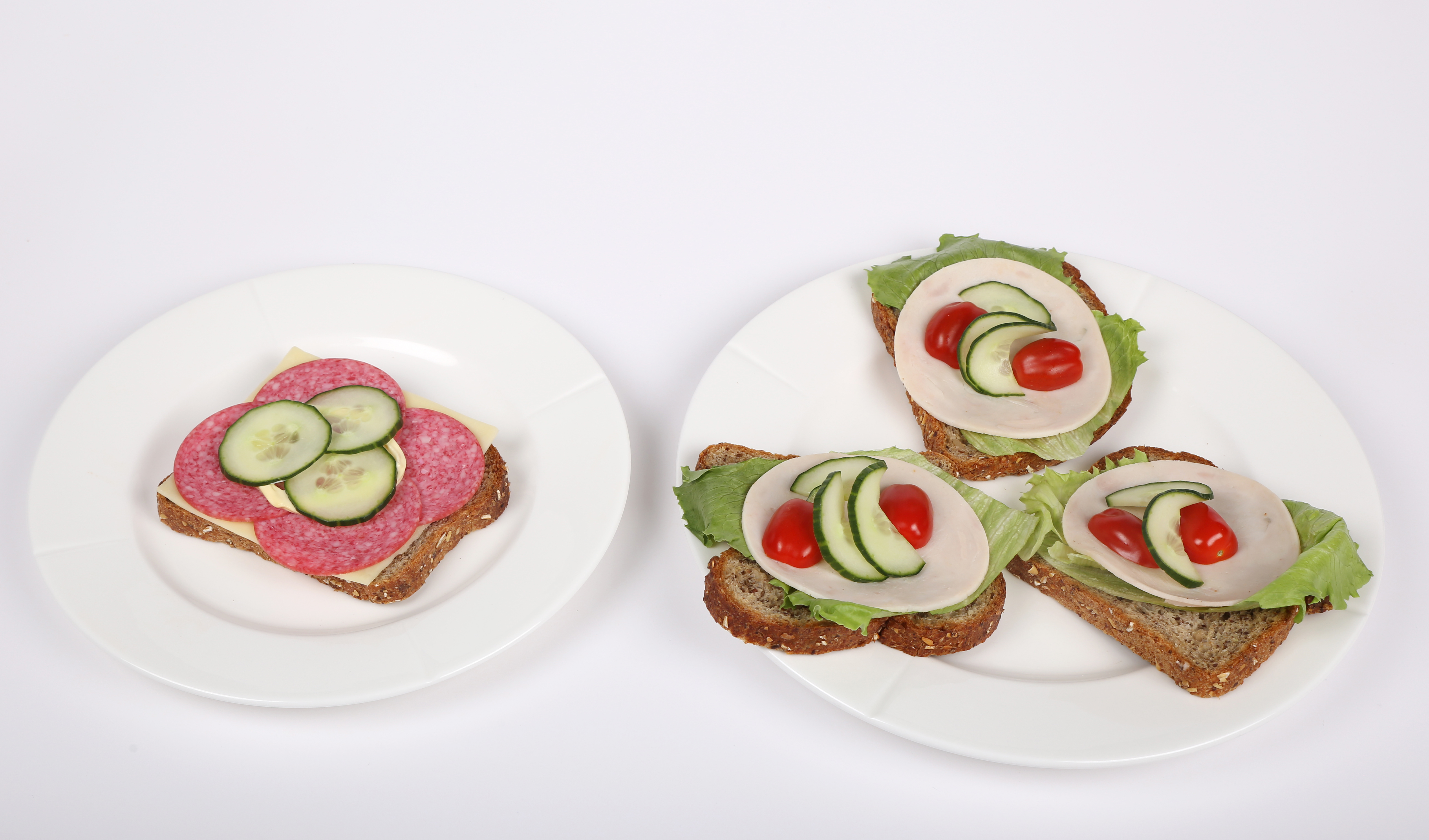 Et fat med en brødskive med majones, salami og agurk, samt et fat med tre brødskiver med salatblad, kalkunpålegg, tomater og agurkskiver.