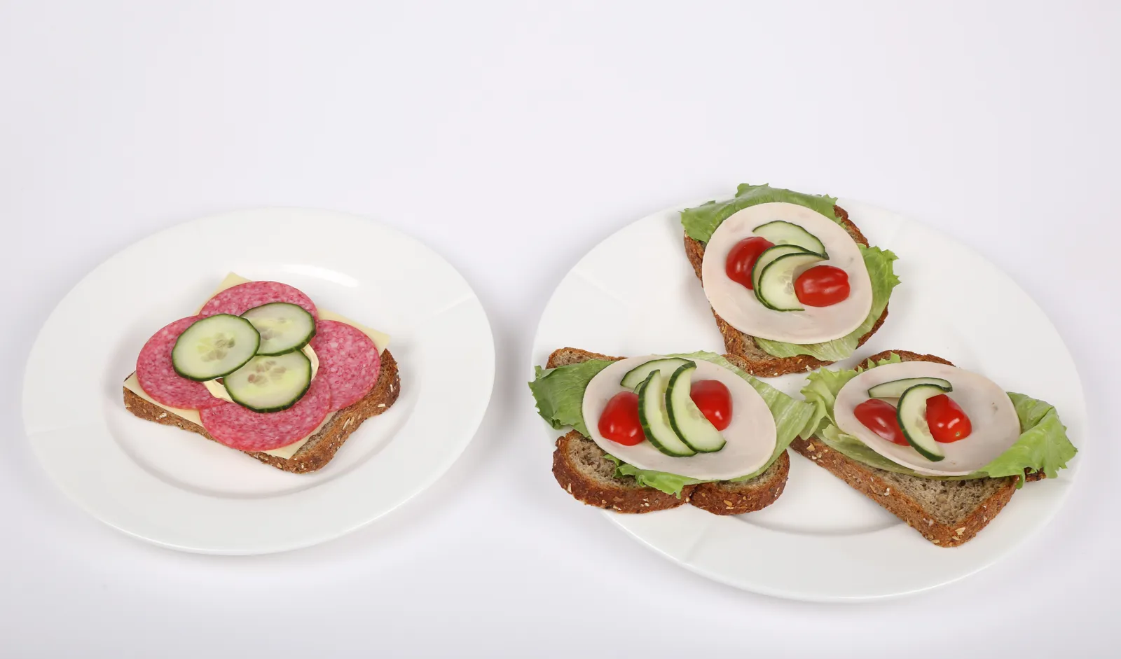 Et fat med en brødskive med majones, salami og agurk, samt et fat med tre brødskiver med salatblad, kalkunpålegg, tomater og agurkskiver.