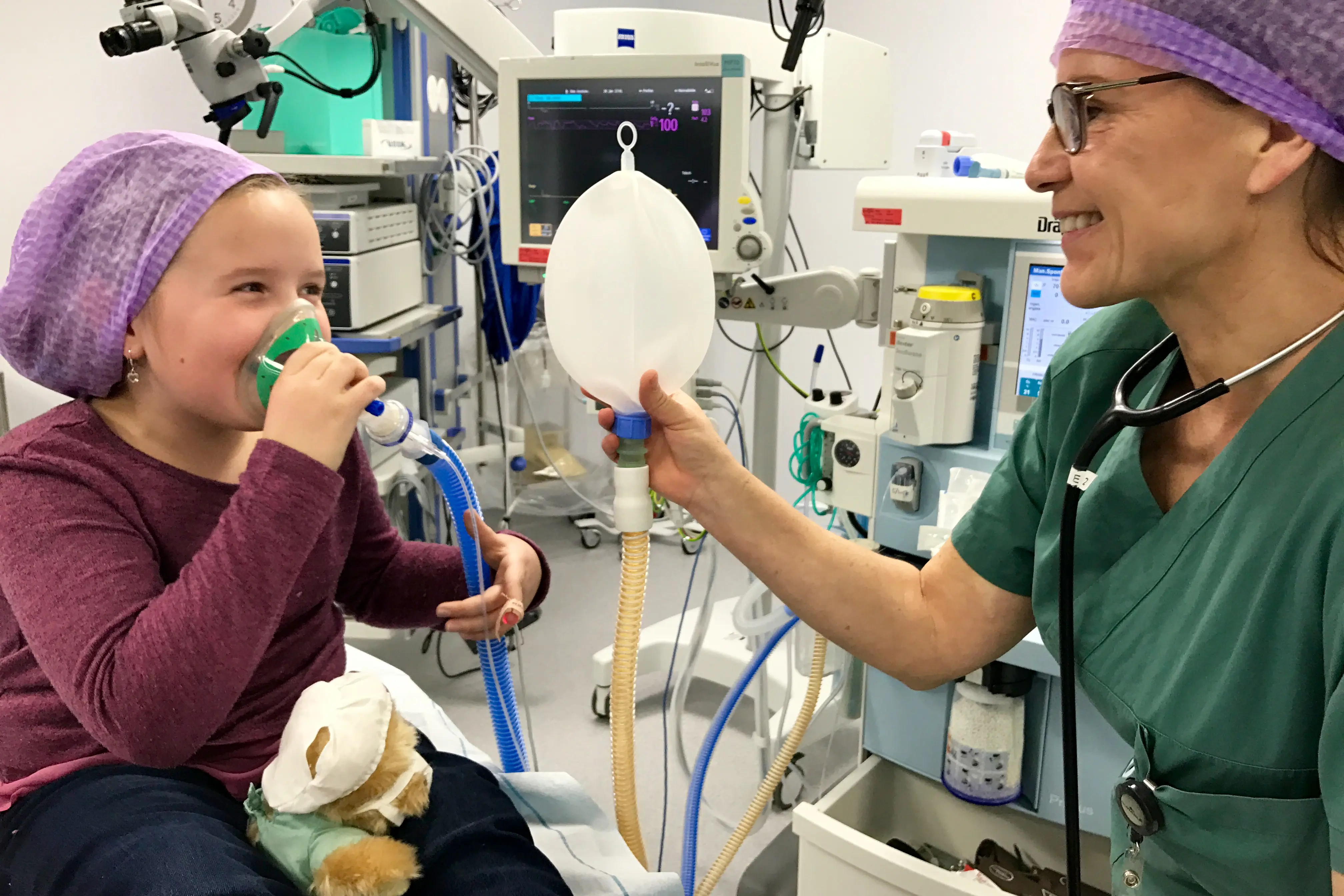 Ei jente på operasjonsstova pustar i ei maske. Anestesisjuk held ein ballong som ein blåser opp når ein pustar i maska. 