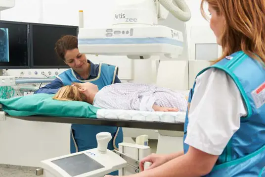 En jente ligger i et røntgenapparat, mens mor og radiograf sitter på hver sin side